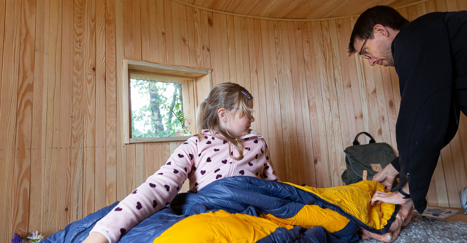 En flicka ligger i en sovsäck i ett vindskydd och pappan hjälper henne.